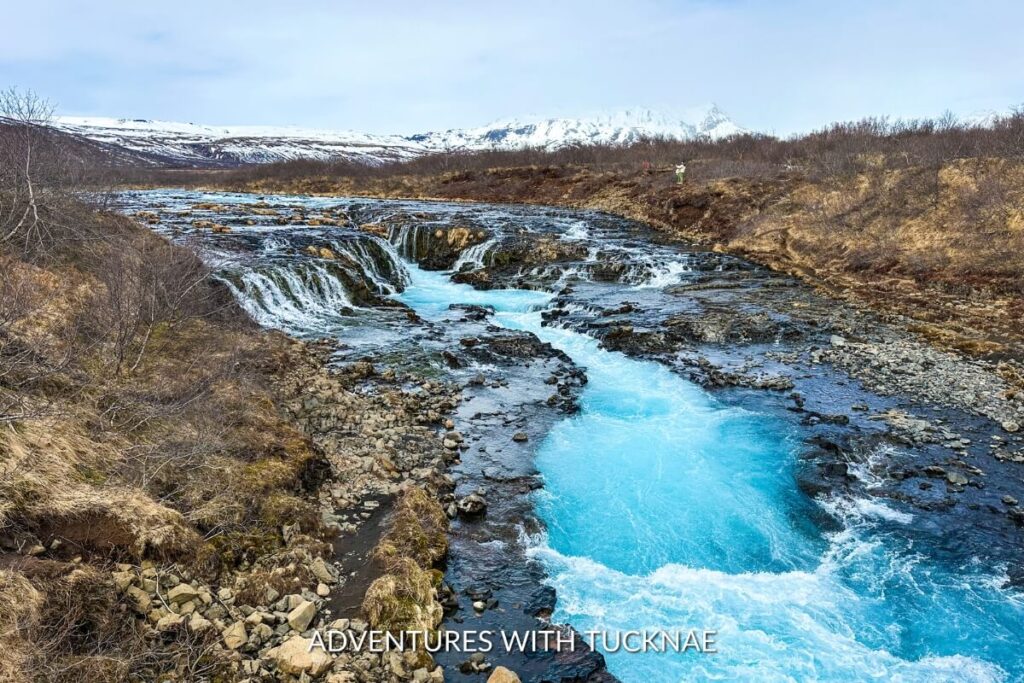 A distant view of Brúarfoss waterfall showing its vibrant blue waters cascading through a rocky riverbed, surrounded by sparse winter vegetation.