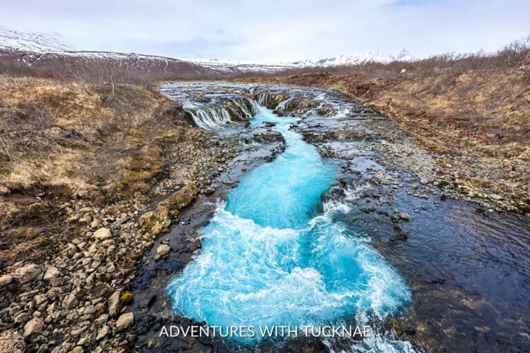 Vivid image of the turquoise waters of Brúarfoss waterfall cascading through a rugged landscape, with snow-dusted mountains in the background.