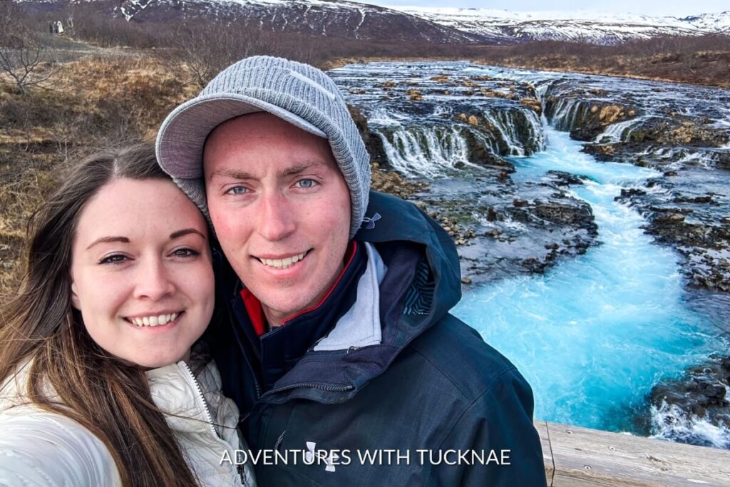 Tucker and Janae posing for a selfie with Brúarfoss waterfall's mesmerizing turquoise waters and rugged terrain in the background.