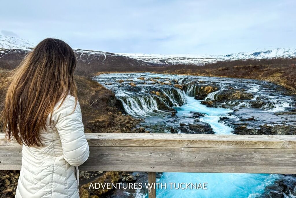 Janae looking out over Brúarfoss waterfall, framed by its stunning blue waters and the snow-capped mountains in the distance.
