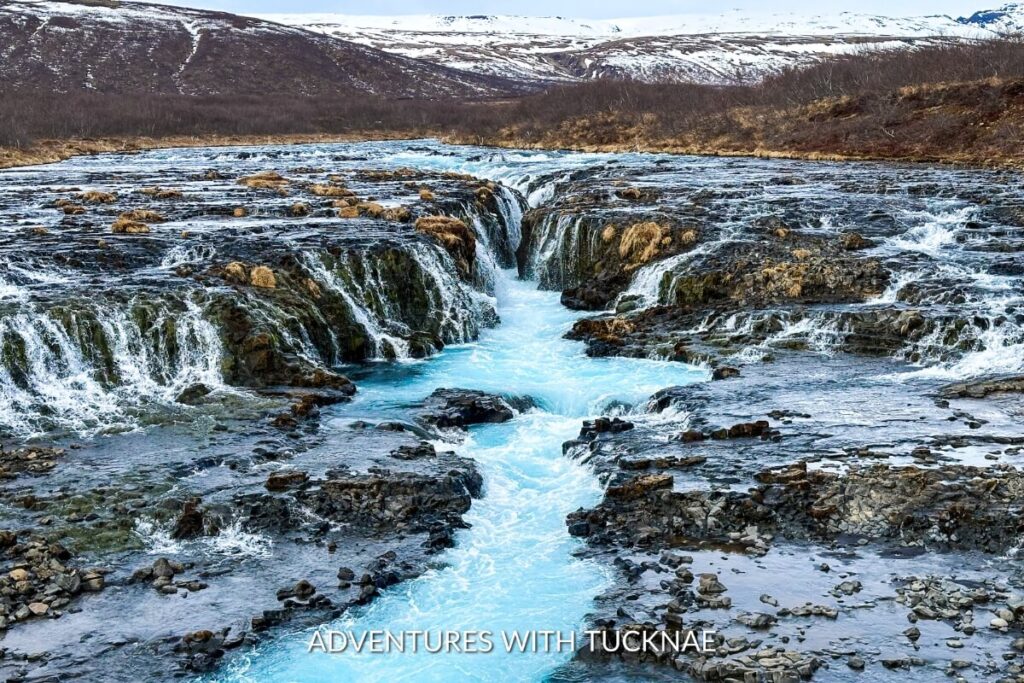 Wide-angle view of the Brúarfoss waterfall showcasing a series of small cascades with bright blue glacial waters flowing through a rocky riverbed, framed by a barren landscape and distant snow-covered hills.