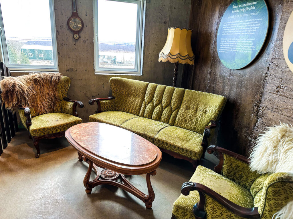 Cozy waiting area at Efstidalur II featuring vintage-style green upholstered seating with ornate wooden arms and backs, paired with a round marble-top coffee table. The rustic charm is accentuated by a furry throw over one chair, a pendulum clock on the concrete wall, and a traditional lampshade. A large information board about the farm's history hangs on the wood-paneled wall, enhancing the quaint, historical atmosphere of the space.