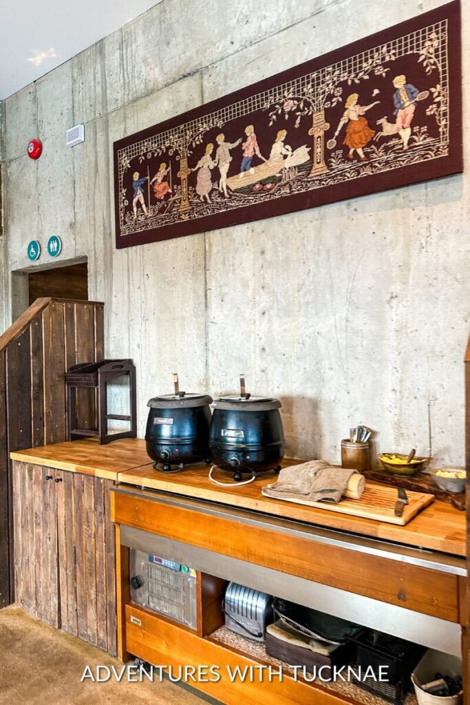 A rustic soup bar setup inside a restaurant, featuring two large black pots on a wooden counter, with a tapestry depicting traditional scenes hanging above.