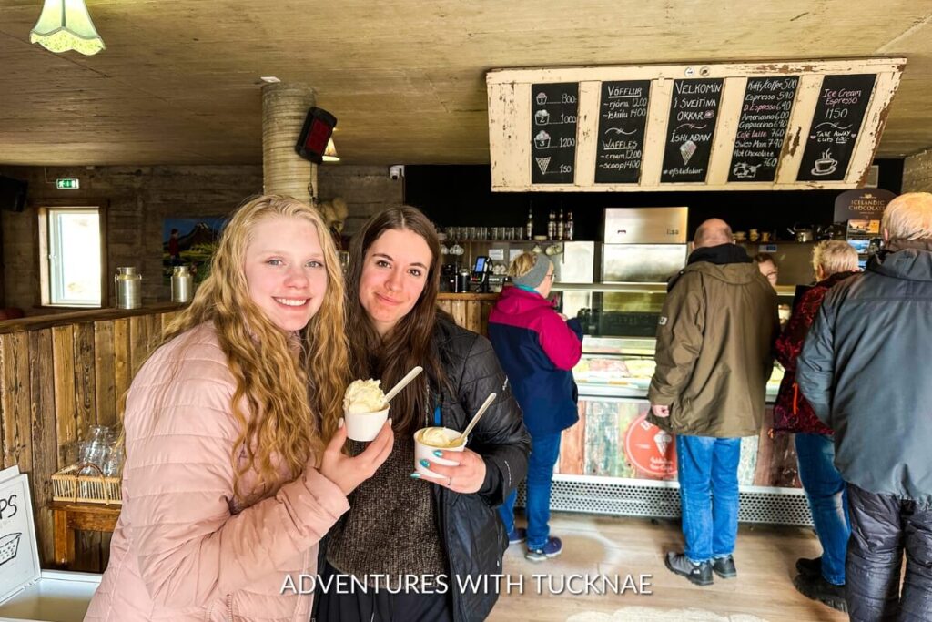 Bree and Alyssa enjoying ice cream inside a rustic farm shop, with a smile, surrounded by other patrons and a vintage-style ice cream menu board in the background.