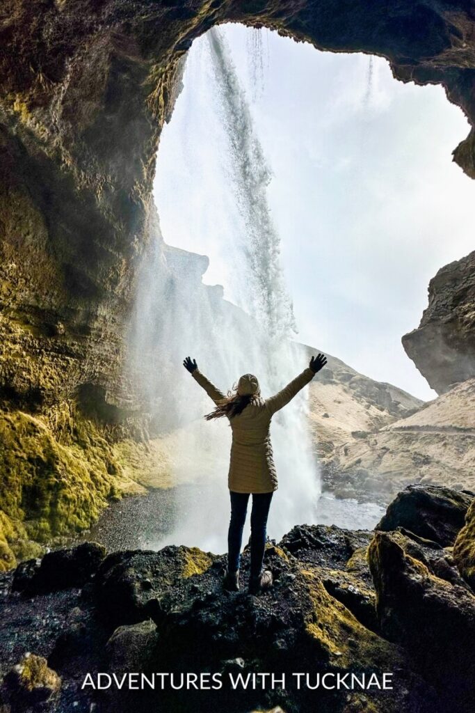 A girl standing behind Kvernufoss waterfall with her hands stretched up to the sky. 