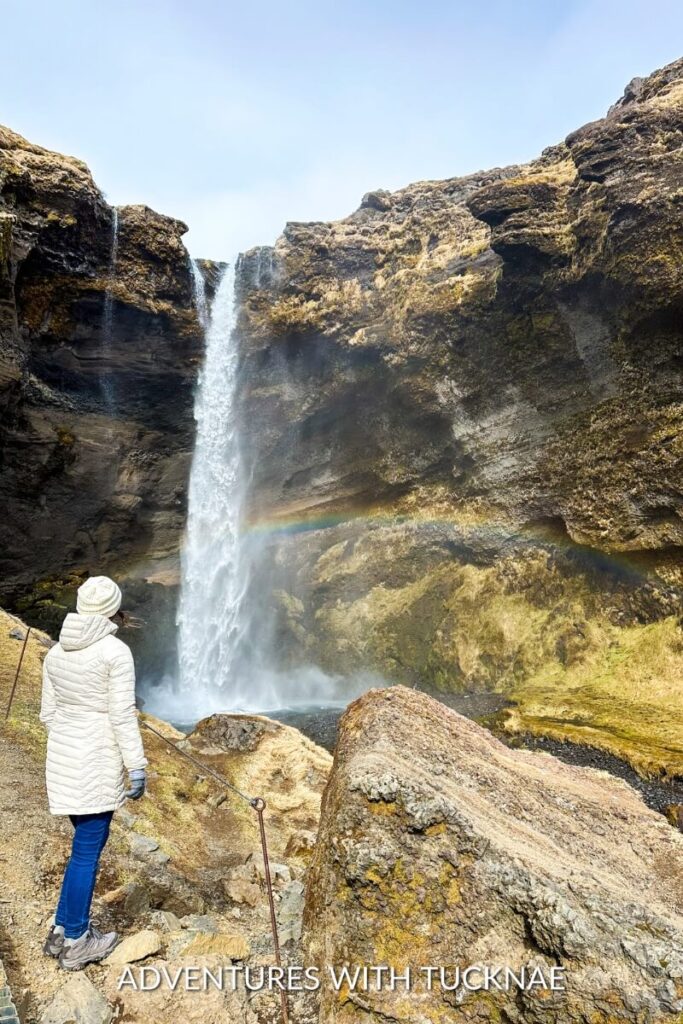 A girl standing in front of and facing the beautiful Kvernufoss waterfall in Iceland with a rainbow in the mist from the falls.