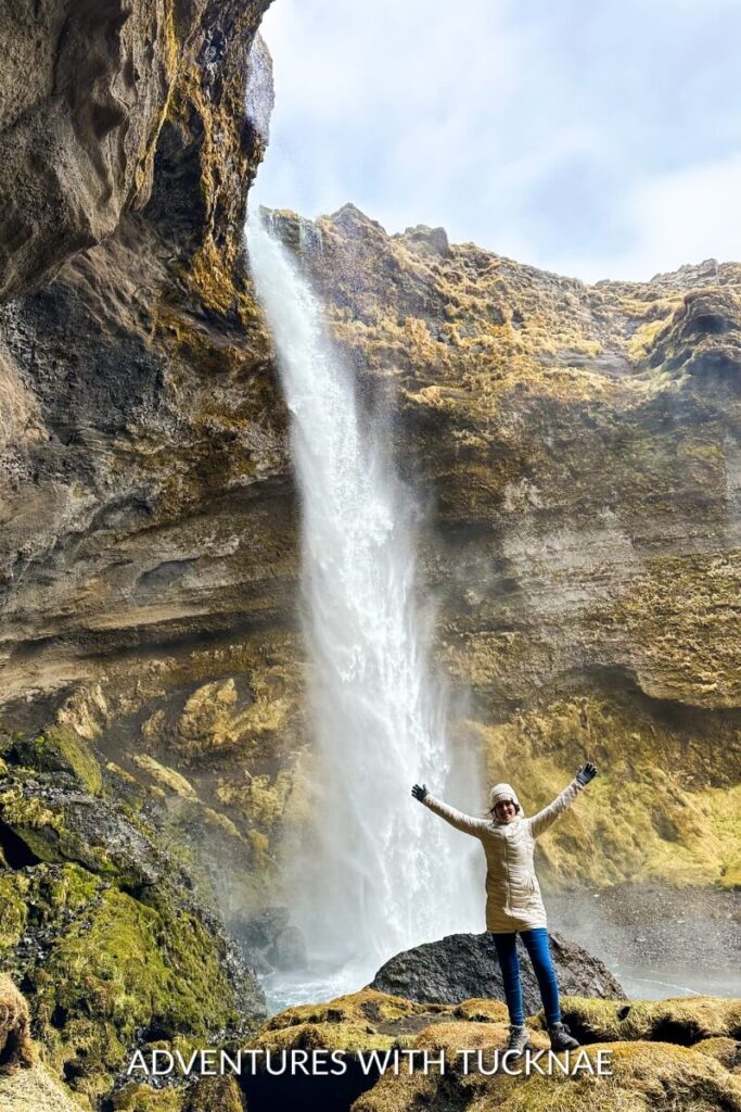 A girl standing with her arms up in front of Kvernufoss waterfall in Iceland. 