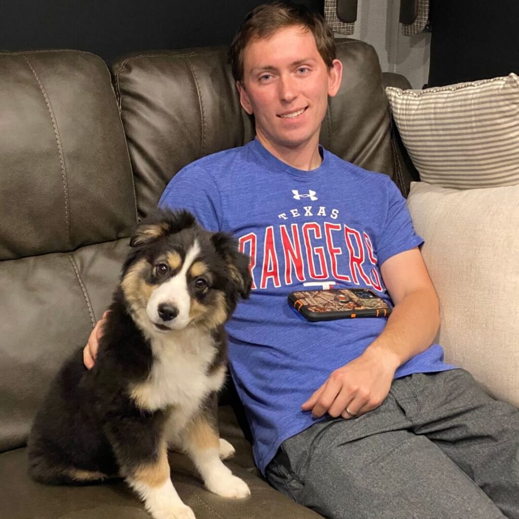 Tucker sits on a couch with Marvel, the Tri-Color Mini Aussie puppy, who is looking up with a curious expression.