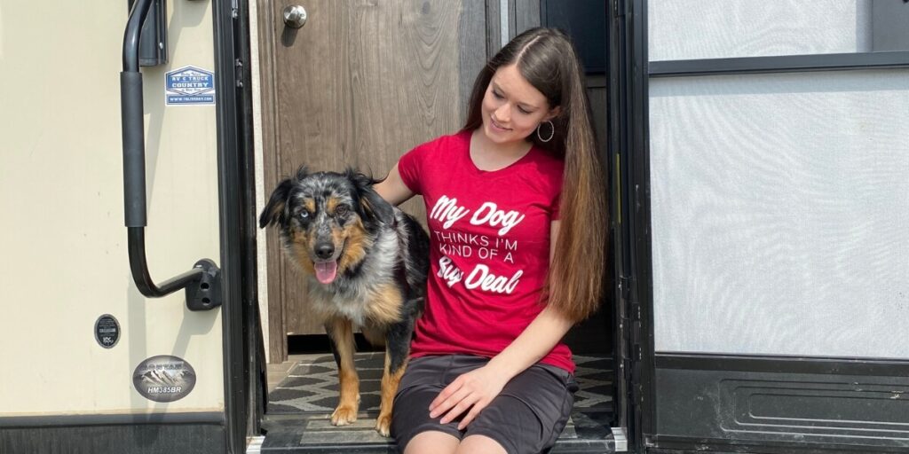 Janae sits on the steps of an RV with her Blue Merle Mini Aussie, Cap, who is panting and standing beside her. She is wearing a red shirt that says "My Dog Thinks I'm Kind of a Big Deal."