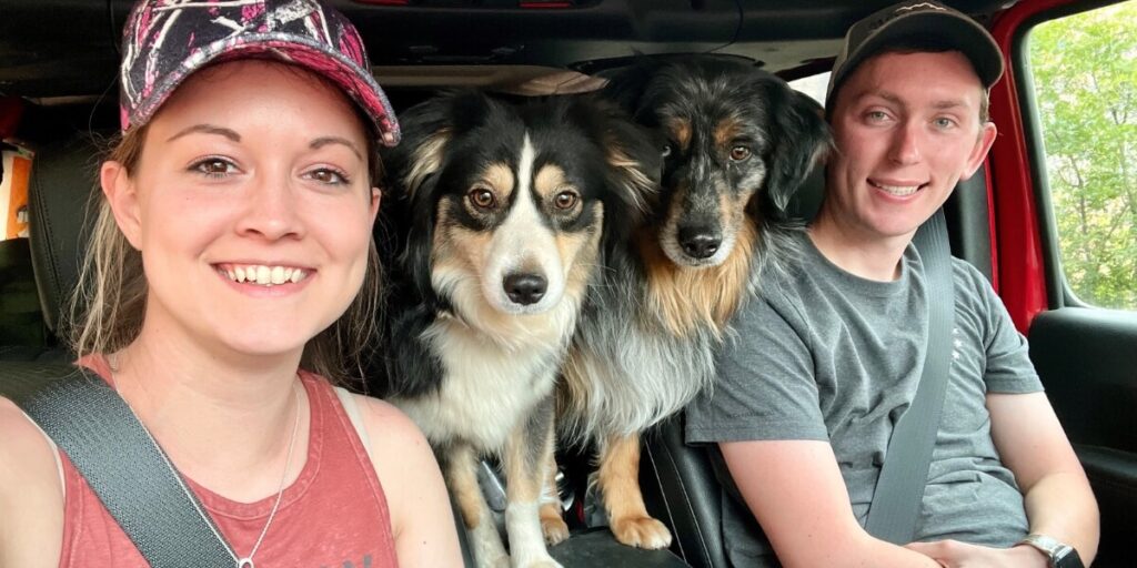 Janae and Tucker smile for a selfie inside a vehicle, accompanied by their two dogs, Cap and Marvel, who are sitting between them.