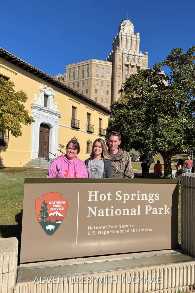 Tucker, Janae, and another person standing by the entrance sign of Hot Springs National Park with historic buildings in the background.