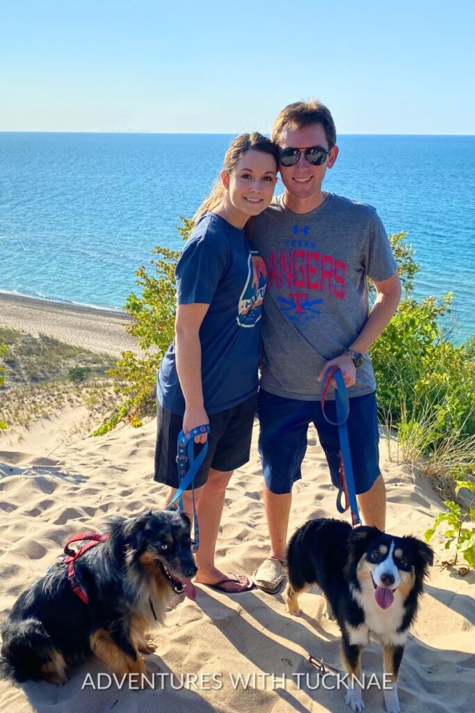 Tucker and Janae standing on a sandy dune overlooking a lake, accompanied by Cap and Marvel at Indiana Dunes National Park.