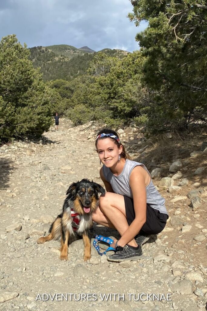 Janae and Cap pose on a rocky hiking trail surrounded by trees and mountains, with Janae kneeling beside Cap.