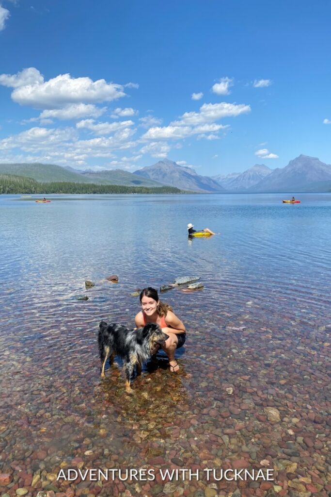 Janae kneels in shallow water with Cap at a lake, while people kayak and float in the background under a clear blue sky.