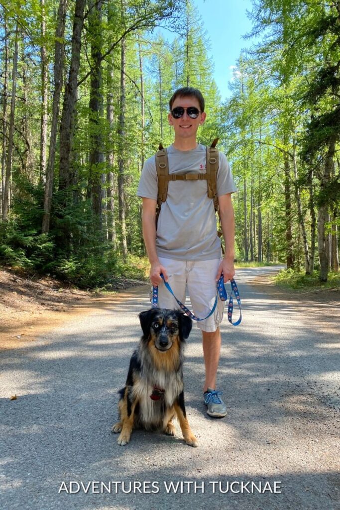 Tucker and Cap take a break on a forest path in Glacier National Park, with Tucker holding Cap's leash and both enjoying the shade.