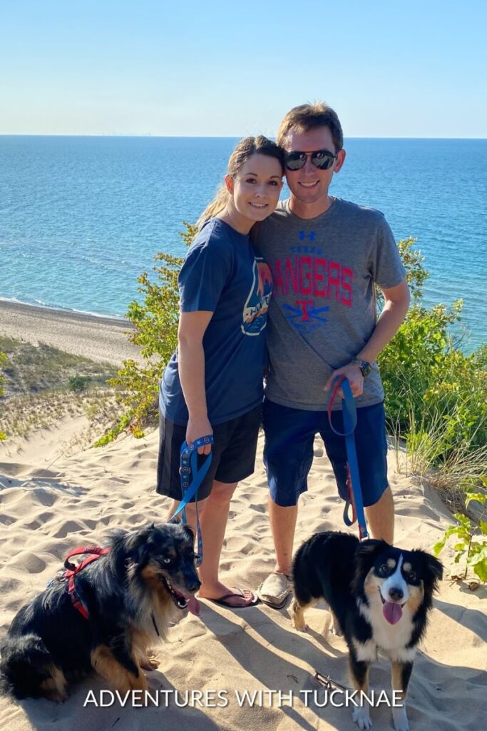 Janae and Tucker smile while standing on a sandy dune overlooking a beautiful ocean, with Cap and Marvel sitting beside them on leashes.