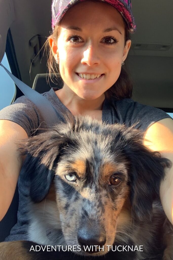 Janae smiles while holding Cap, a blue merle dog, on her lap inside a vehicle.