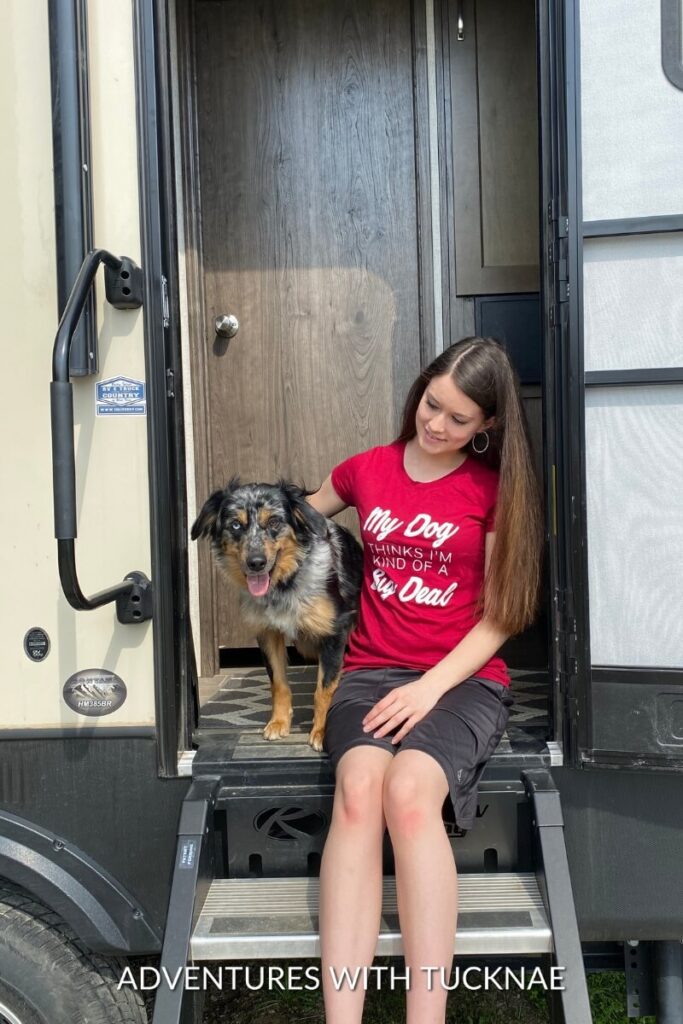 Janae sits on the steps of an RV with Cap beside her.