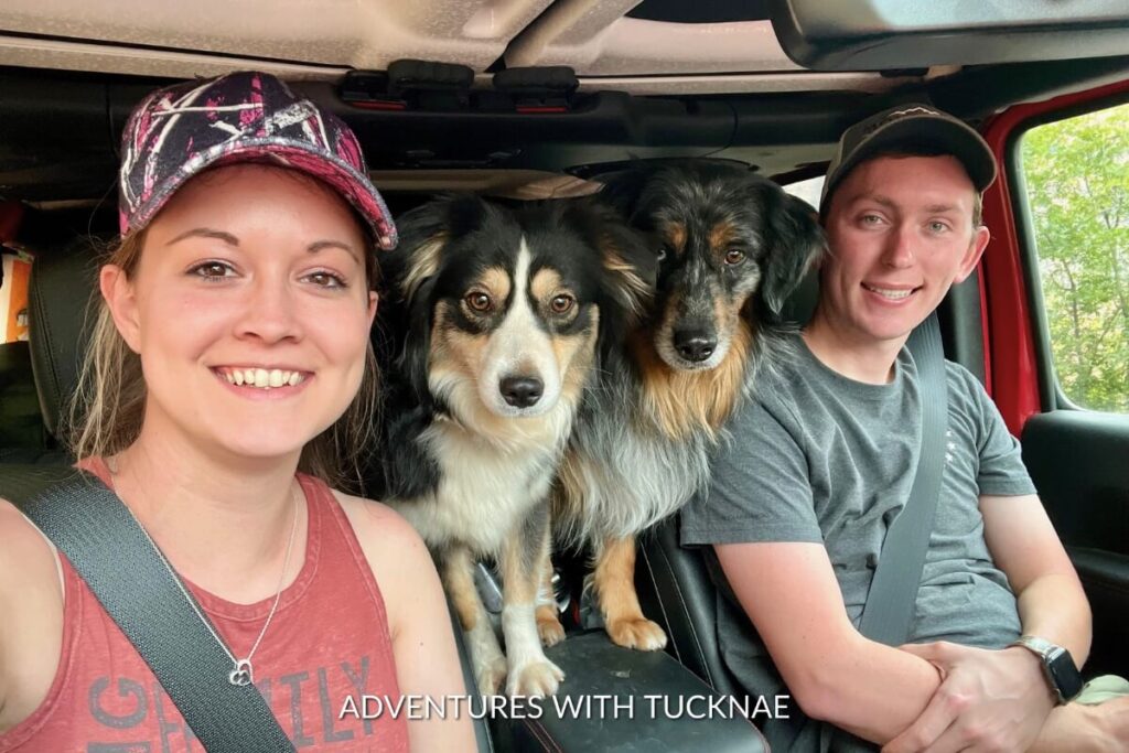 Janae and Tucker smile for a selfie inside a vehicle, accompanied by their two dogs, Cap and Marvel, who are sitting between them.