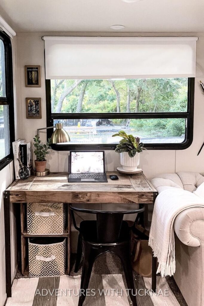 A rustic RV office setup with a wooden desk, metal chair, and a laptop. The space is decorated with small plants, a desk lamp, and framed artwork, providing a warm and functional workspace. The window overlooks lush greenery, creating an inviting RV office environment.