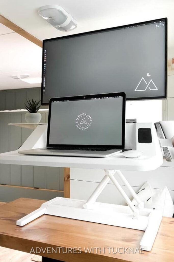 A modern RV office setup featuring a dual-monitor setup with a sleek standing desk. The setup includes a large monitor mounted above a laptop, both displaying a minimalist mountain logo. This compact design maximizes productivity in small spaces, ideal for travel trailer office ideas.