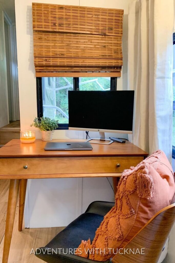 A simple RV office space featuring a classic wooden desk and a black office chair. The desk includes a single monitor, a laptop, a potted plant, and a lit candle, providing a warm and inviting workspace for those seeking RV office ideas.