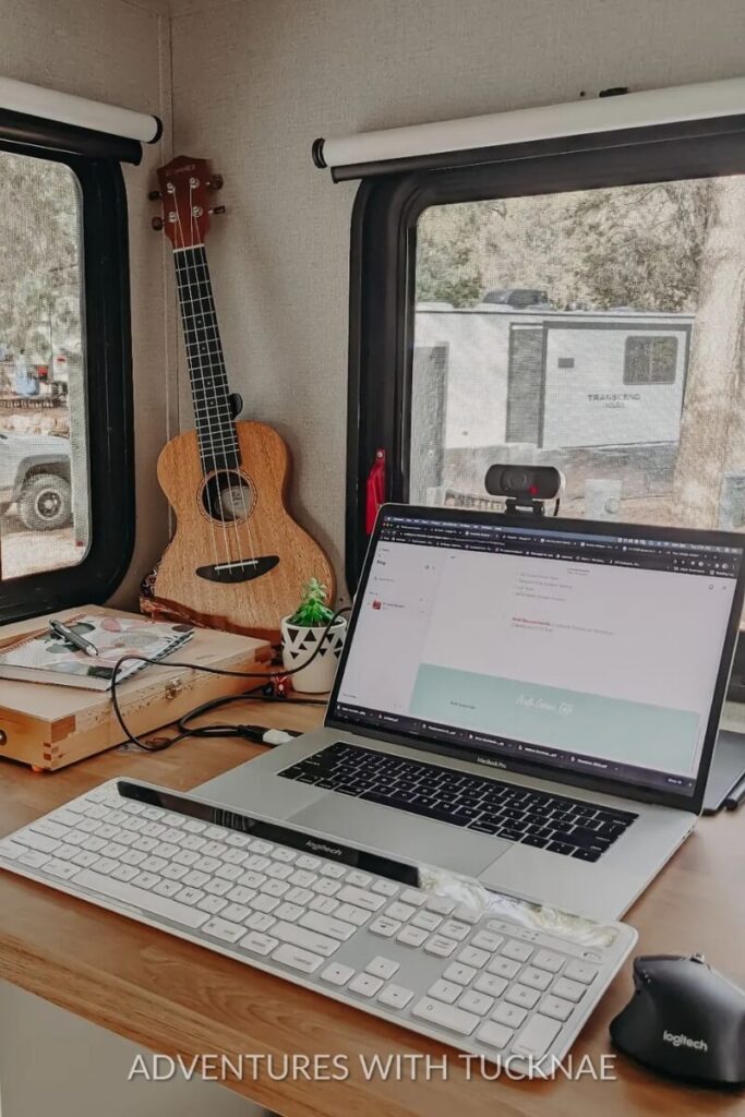 An RV office setup featuring a laptop, external keyboard, and a webcam. A ukulele rests in the corner, adding a personal touch, while the window shows a glimpse of another RV parked outside. This cozy workspace reflects a blend of productivity and creativity in a travel trailer office idea.