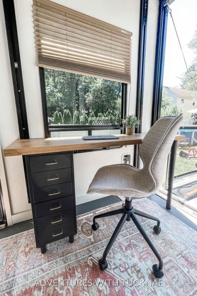 A minimalist RV office with a black filing cabinet, wooden desk, and a grey ergonomic chair. The large window allows natural light to brighten the workspace, offering a simple yet elegant RV office setup.