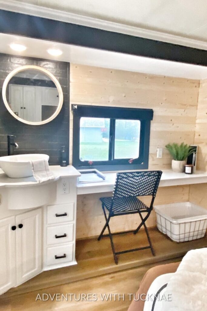 A bright RV office space doubling as a bathroom vanity. A black woven chair sits at a white floating desk near a window, with modern lighting overhead, offering a multi-functional space within the RV.