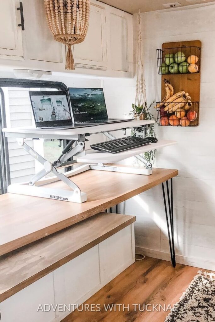 A standing desk setup in an RV kitchen with a dual-laptop configuration. The desktop features a monitor riser and a variety of fresh fruit stored on the wall, blending productivity and kitchen space in this RV office idea.