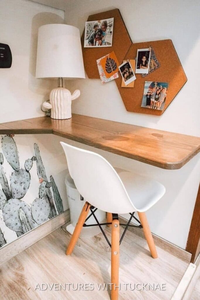 A small RV workspace with a wooden corner desk, a cactus-patterned wallpaper, and a white chair. A lamp and a corkboard with pinned photos personalize the space, ideal for small RV office ideas.