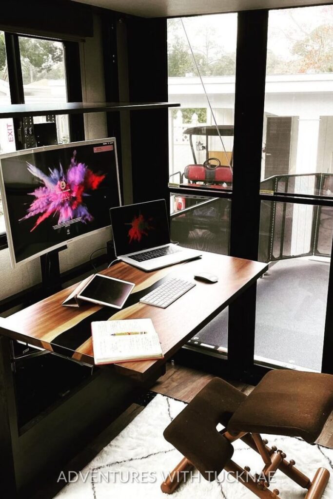 A sleek RV office setup with a large monitor, laptop, and tablet. The workspace is positioned near a window, featuring a modern chair and a wooden desk with a black stripe, creating a tech-friendly and stylish RV office.