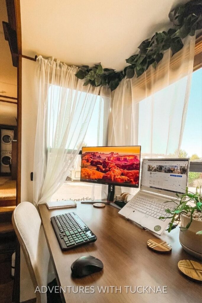 A bright and airy RV office setup with a dual-screen workstation. The desk is framed by sheer white curtains and faux greenery, adding a serene ambiance to the workspace, which is ideal for an RV or travel trailer office setup.