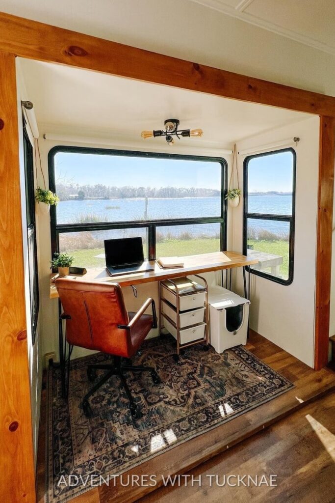 A cozy RV office space with a large window providing a scenic lake view. The setup features a simple wooden desk with a laptop, drawers, and a brown leather chair, offering inspiration for a peaceful and productive RV office setup.