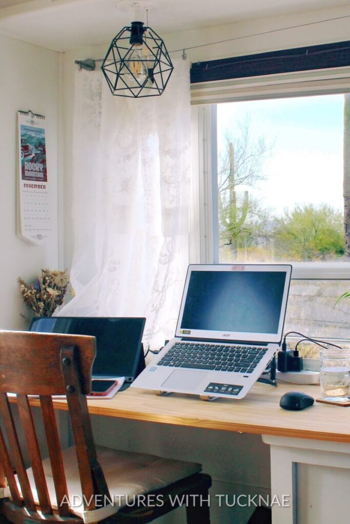 A bright and inviting RV office space with white curtains and a geometric light fixture. Two laptops sit on a wooden desk alongside a glass of water, showing a comfortable and organized workspace perfect for a travel trailer office setup.