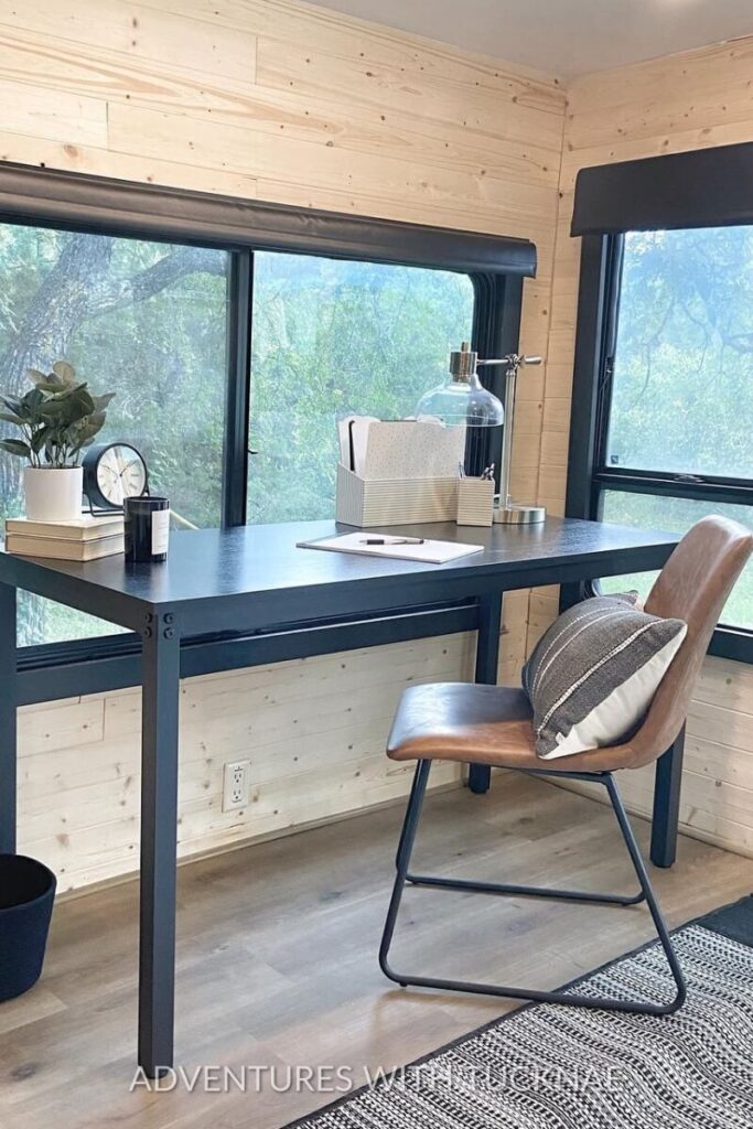 A rustic RV office setup with a black desk and a tan leather chair, set against large windows that overlook a forested area. The desk includes a few essentials like a plant, a clock, and some neatly organized paperwork, demonstrating an ideal RV office environment.