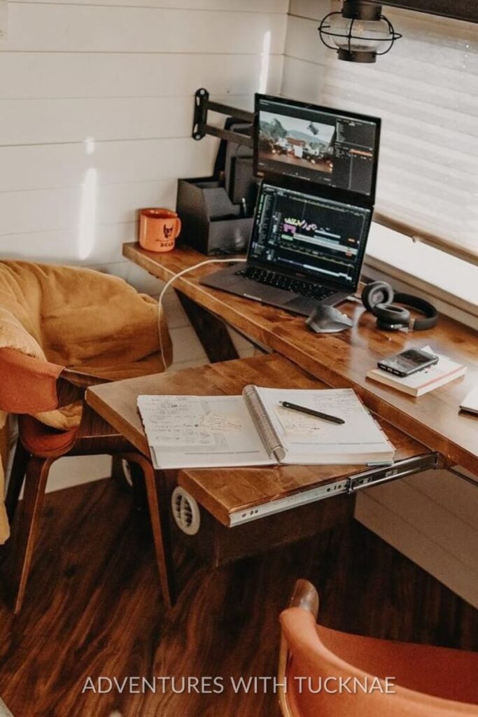 A compact yet cozy RV office featuring a wooden desk with a notebook and headphones. Two monitors are mounted, showing video editing software, offering inspiration for a creative workspace within an RV.