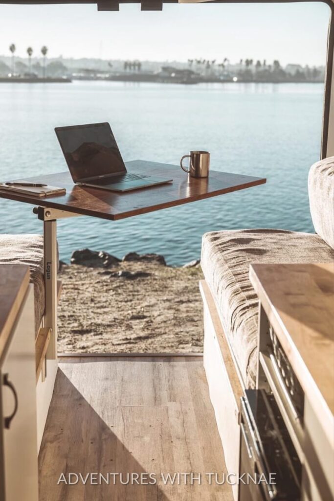 A minimalist RV workspace with a foldable wooden desk that overlooks a scenic waterfront view. The desk includes a laptop, a mug, and a pen, showcasing an inspiring environment for remote work while traveling in an RV.