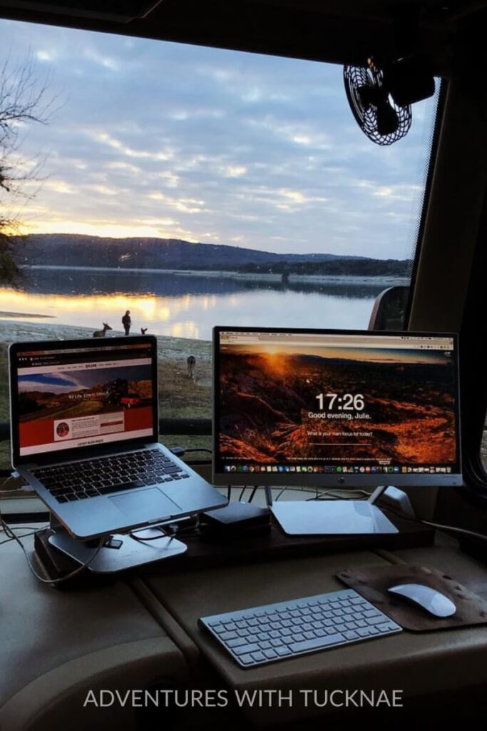 A dual-monitor RV office setup with a panoramic lake view through the windows. The desktop shows productivity apps on both screens, while a keyboard and mouse complete the mobile office configuration, ideal for digital nomads.