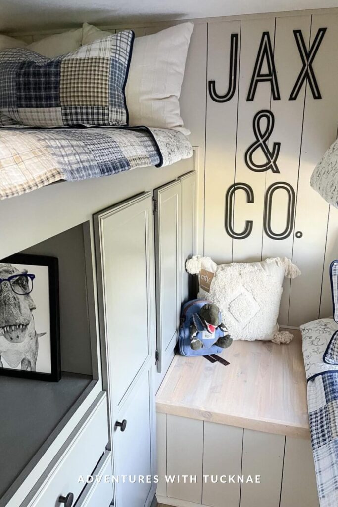A compact RV bunk area with a cozy top bunk bed dressed in plaid bedding, built-in cabinets below, and a decorative wall reading “JAX & CO.” A plush white pillow, a small backpack with a stuffed animal, and a framed illustration of a glasses-wearing T-Rex add playful charm.