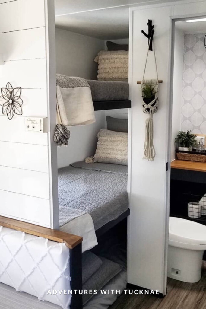 Minimalist RV bunk beds with gray bedding and off-white tasseled throw pillows, tucked beside a bathroom nook. The decor includes a macrame plant hanger and small wooden shelf with baskets and greenery for a natural touch.