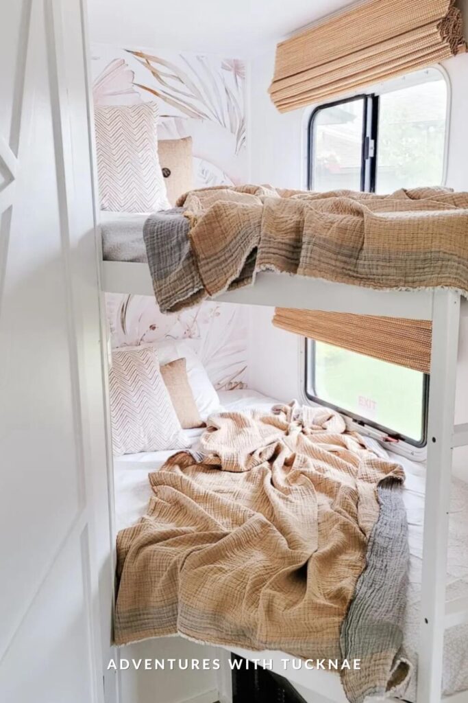 Bright and soft RV bunk space styled with blush-toned bedding and botanical wallpaper. Natural woven blinds and a window bring in daylight, while oversized pillows and cozy blankets invite relaxation.