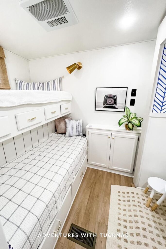 Fresh and modern RV bunk area in all-white tones with subtle black and brass accents. The beds are dressed in checkered and striped bedding, and a framed black-and-white photo and potted plant sit on a small white cabinet.