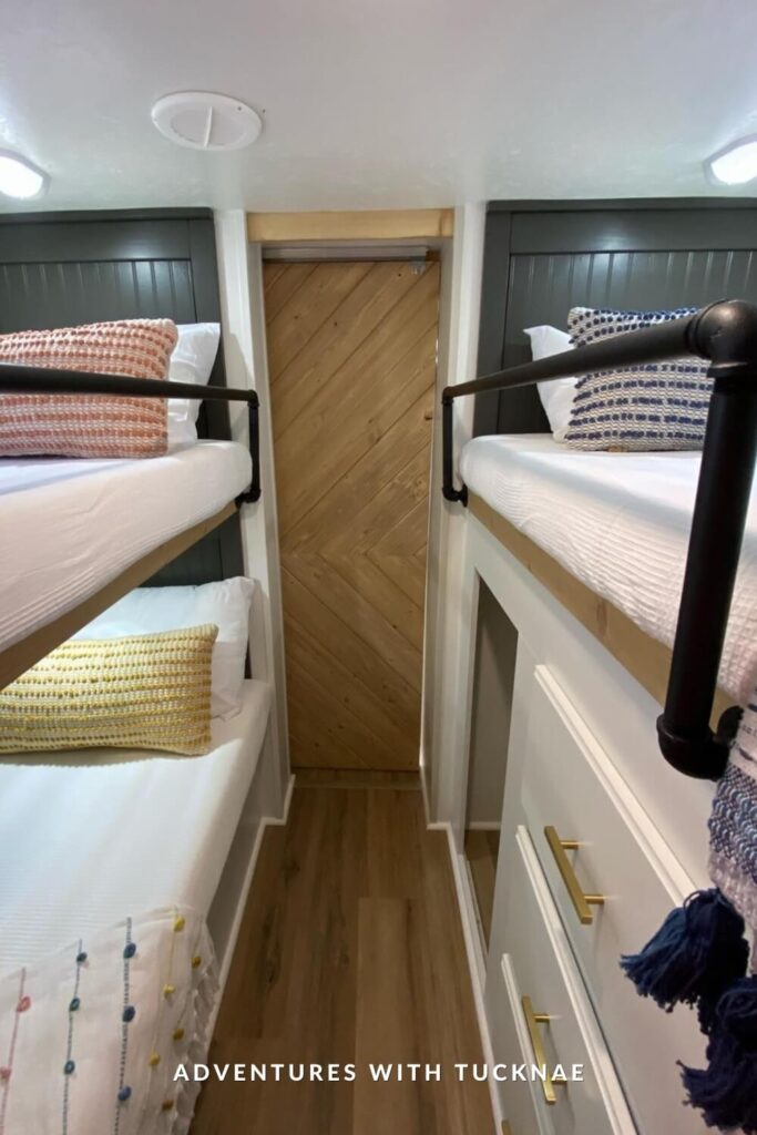 A sleek RV bunk hallway featuring two twin bunk beds with black metal railings, crisp white bedding, and colorful woven throw pillows. A chevron-patterned wood door adds texture and warmth at the far end.
