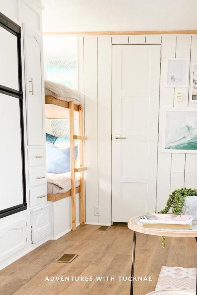 Minimalist surf-style RV space with light wood flooring and a bunk tucked into the wall beside a vertical wood-paneled door. The beds have pale beige bedding, and coastal-themed art in light frames hangs on the wall.