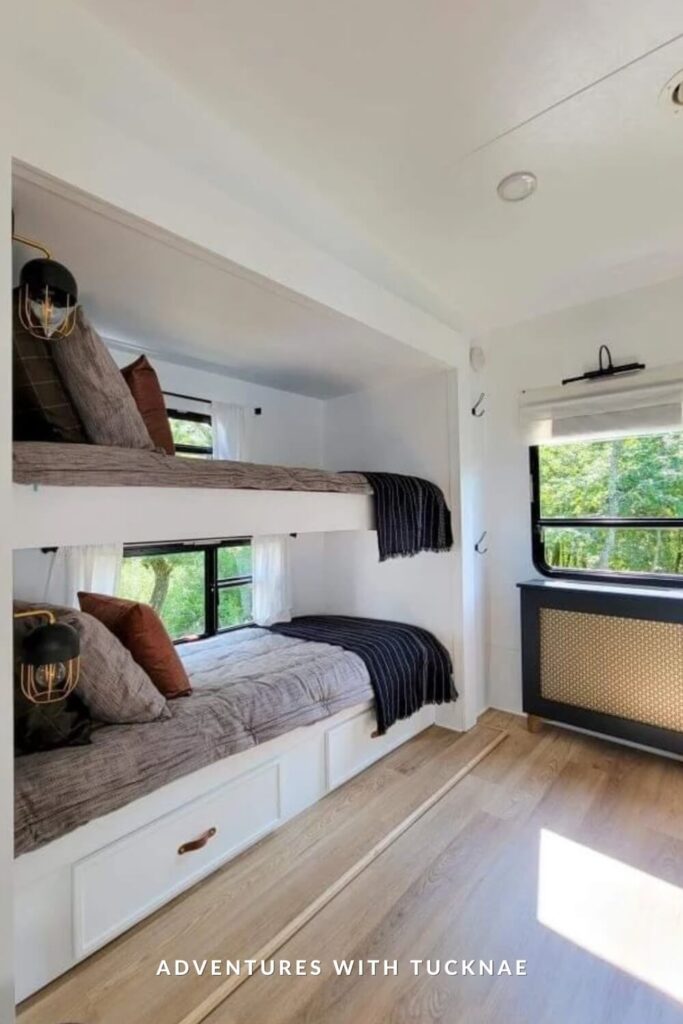 Modern and airy RV bunk nook with white framing and drawers beneath each bed. The bedding is a mix of gray and rust tones, with black industrial-style sconces mounted beside each bunk for reading light.
