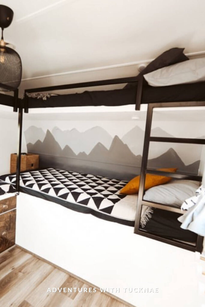 Adventure-themed RV bunk room with black and white geometric bedding and a painted mountain mural across the back wall. Black metal frames define the beds, with a bold mustard pillow as an accent.