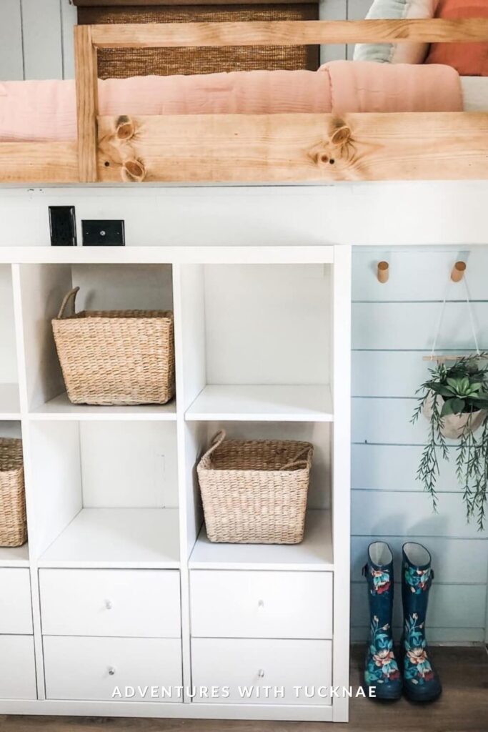 Bright RV bunk setup with natural wood guardrails, soft pink bedding, and a cubby-style white storage unit below. Woven baskets, potted greenery, and a pair of floral-patterned rain boots enhance the fresh and playful aesthetic.