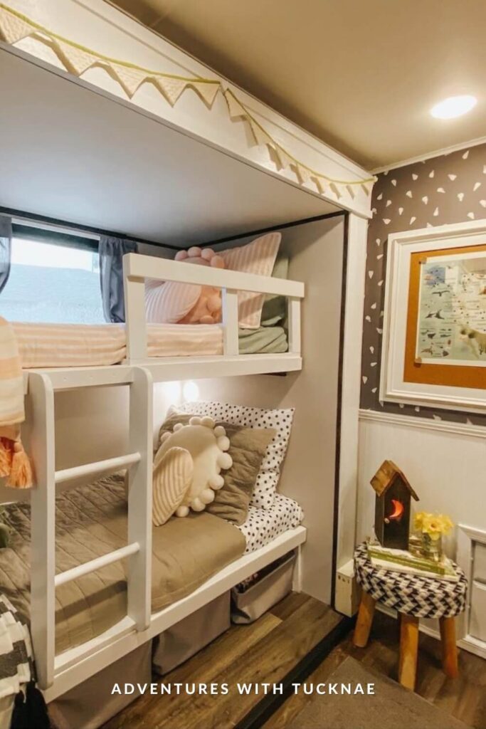 A whimsical RV bunk room with a white bunk bed framed by neutral walls and soft lighting. The bedding includes polka dot and textured pillows, with a fun pom-pom pillow and a nightlight built into the wall. A small side table holds a house-shaped lamp and vase of yellow flowers.