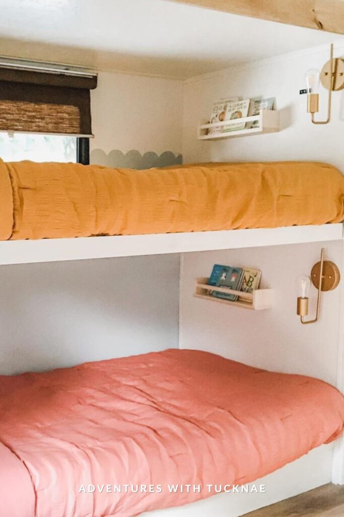 Minimalist and cheerful bunk beds in an RV setup, with a mustard yellow top blanket and coral-toned bottom blanket. Small floating bookshelves and brass wall sconces are mounted next to each bunk, creating a cozy reading-friendly space.